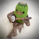 A green plush toy character with brown limbs sitting on a small wooden stool and playing a banjo, photographed against a soft light gray background.