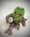 A green plush toy character with brown limbs sitting on a small wooden stool and playing a banjo, photographed against a soft light gray background.