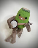 A green plush toy character with brown limbs sitting on a small wooden stool and playing a banjo, photographed against a soft light gray background.