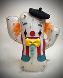 A cactus-shaped vinyl plush dressed like a clown with silver metal spikes, a black beret, orange and black hair accents, a red and white striped vest, a yellow bow tie, and turquoise details, shown on a neutral gray background.