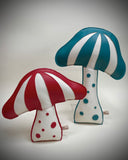 Two mushroom-shaped decorative objects on a light gray studio background. One has red and white stripes with red polka dots on the stem, and the other has teal and white stripes with teal polka dots on the stem.
