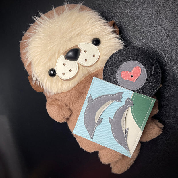 Plush sculpture of a brown sea otter with a cute cartoonish fluffy face. It holds a record sleeve with 2 dolphins on it and a heart shaped record comes out of it.