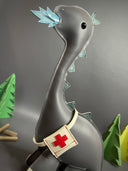 Vinyl plush sculpture of a mostly flat dimension long neck dinosaur, gray in color and with blue flames coming out of its mouth. It has a first aid kit tied around its body.