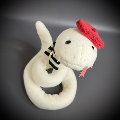 Off white plush sculpture of a snake with 2 small fangs and a pink pleather tongue, positioned out of its closed mouth. It wears a black and white striped scarf and a red beret.