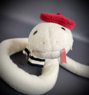 Off white plush sculpture of a snake with 2 small fangs and a pink pleather tongue, positioned out of its closed mouth. It wears a black and white striped scarf and a red beret.