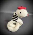Off white plush sculpture of a snake with 2 small fangs and a pink pleather tongue, positioned out of its closed mouth. It wears a black and white striped scarf and a red beret.