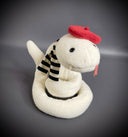 Off white plush sculpture of a snake with 2 small fangs and a pink pleather tongue, positioned out of its closed mouth. It wears a black and white striped scarf and a red beret.
