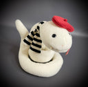 Off white plush sculpture of a snake with 2 small fangs and a pink pleather tongue, positioned out of its closed mouth. It wears a black and white striped scarf and a red beret.