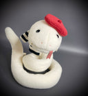 Off white plush sculpture of a snake with 2 small fangs and a pink pleather tongue, positioned out of its closed mouth. It wears a black and white striped scarf and a red beret.