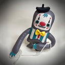 A handmade plush toy sloth, dressed as a clown, featuring a dark gray body, white face, red nose, yellow bow tie, and teal accents, sitting on a light gray background.