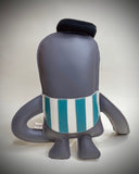 A handmade plush toy sloth, dressed as a clown, featuring a dark gray body, white face, red nose, yellow bow tie, and teal accents, sitting on a light gray background.