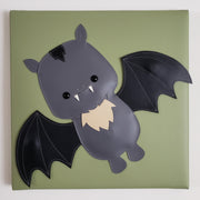 Vinyl canvas flat sculpture of a cartoon style bat, with its wings spread out. It is on a olive green square canvas.