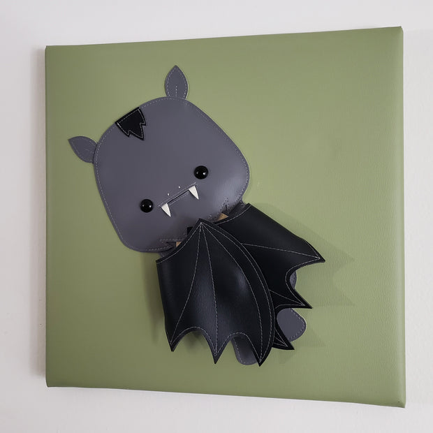 Vinyl canvas flat sculpture of a cartoon style bat, with its wing pulled into its body. It is on a olive green square canvas.