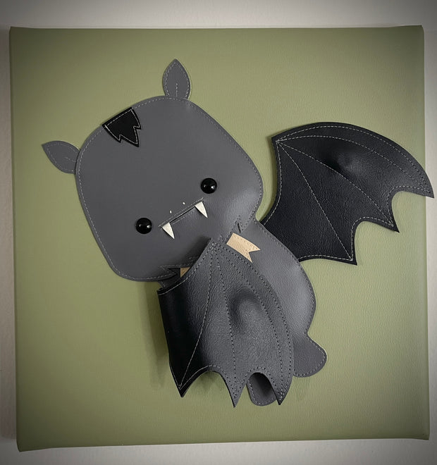 Vinyl canvas flat sculpture of a cartoon style bat, with one wing out and one wing pulled into its body. It is on a olive green square canvas.