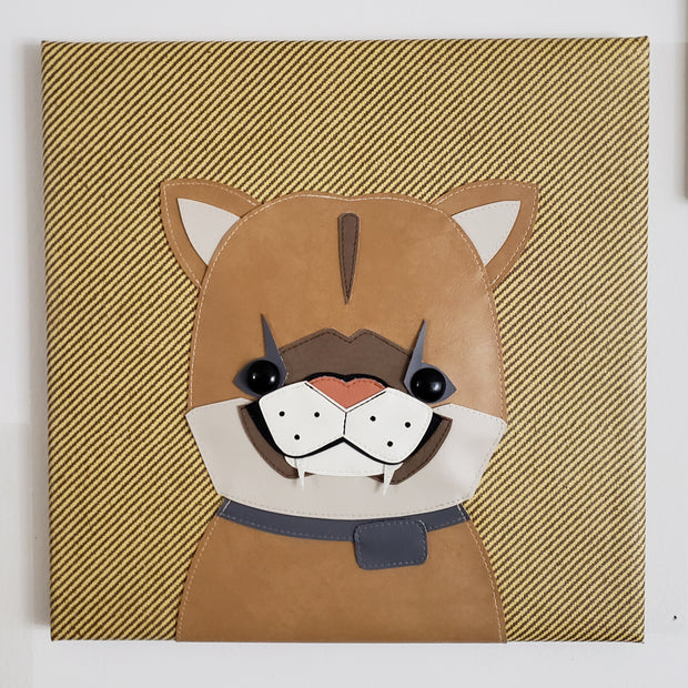 Vinyl fabric sculpture on a flat striped tan square canvas of a cartoon style baby mountain lion, with only its upper body visible.