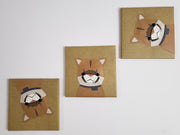 3 square canvases wrapped in tan striped fabric. Atop of each is a brown cartoon style flat sculpture of a mountain lion, each one facing in a different direction.