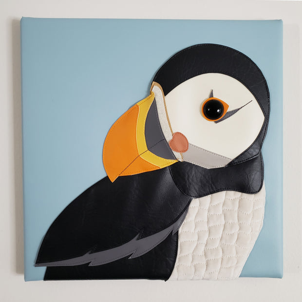 Vinyl fabric sculpture on a flat baby blue square canvas of a cartoon style puffin, looking over its shoulder.