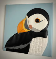 Vinyl fabric sculpture on a flat baby blue square canvas of a cartoon style puffin, looking over its shoulder.