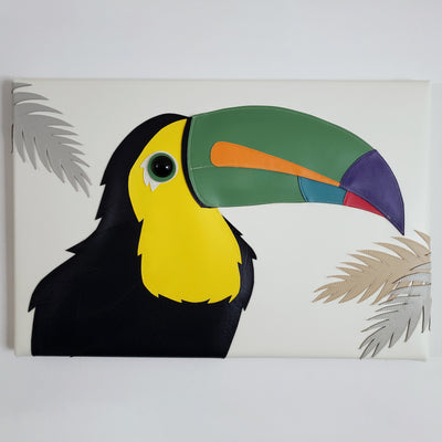 Vinyl canvas flat sculpture of a cartoon style toucan, facing to the right. It is on a horizontal rectangle canvas, white with palm tree leaves coming in from the edges of the canvas.