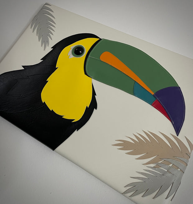 Vinyl canvas flat sculpture of a cartoon style toucan, facing to the right. It is on a horizontal rectangle canvas, white with palm tree leaves coming in from the edges of the canvas.