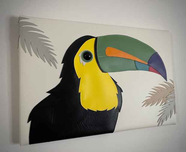 Vinyl canvas flat sculpture of a cartoon style toucan, facing to the right. It is on a horizontal rectangle canvas, white with palm tree leaves coming in from the edges of the canvas.