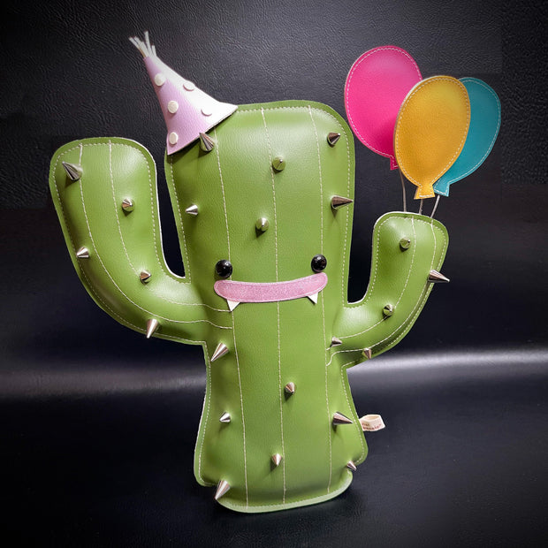 Pleather plush sculpture of a smiling cartoon style cactus, with metal spikes all over its body. It wears a pointed birthday cap and holds 3 balloons in its hand.