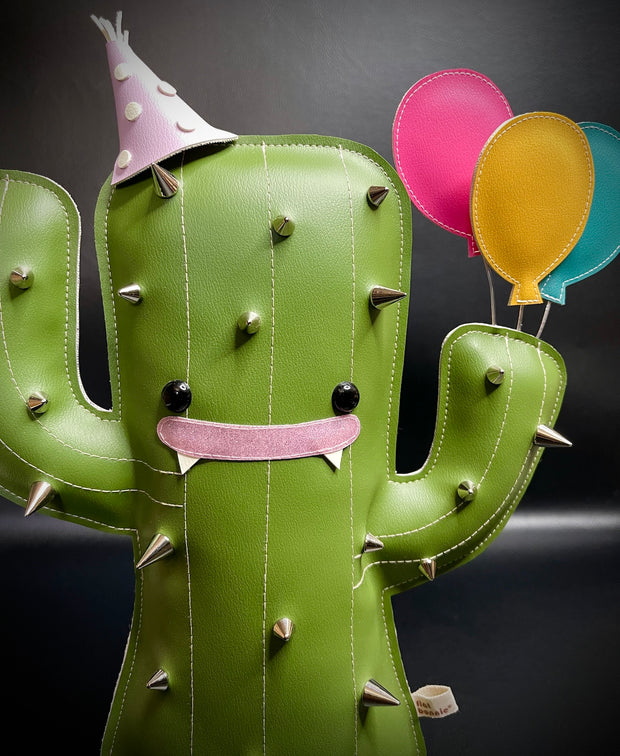 Pleather plush sculpture of a smiling cartoon style cactus, with metal spikes all over its body. It wears a pointed birthday cap and holds 3 balloons in its hand.