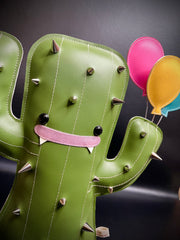 Pleather plush sculpture of a smiling cartoon style cactus, with metal spikes all over its body. It wears a pointed birthday cap and holds 3 balloons in its hand.