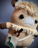 Plush doll of an anthropomorphic highland cow with a furry head and collar. It has a satchel it wears.