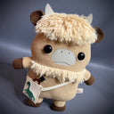 Plush doll of an anthropomorphic highland cow with a furry head and collar. It has a satchel it wears.