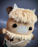 Plush doll of an anthropomorphic highland cow with a furry head and collar. It has a satchel it wears.