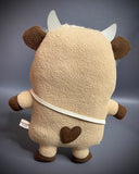Plush doll of an anthropomorphic highland cow with a furry head and collar. It has a satchel it wears.