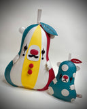 Two plush, bear pear-shaped plush sculptures. Plush on the left is vertically oriented with teal, yellow, white, and red panels, featuring a mustache and red buttons.