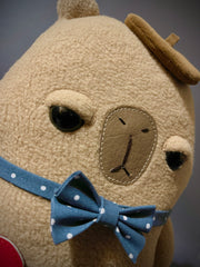 Flat plush sculpture of a capybara with an apathetic expression. It stands on 2 legs like a person and wears a brown beret, a blue bowtie with white polka dots and holds a torn heart.