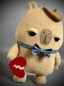 Flat plush sculpture of a capybara with an apathetic expression. It stands on 2 legs like a person and wears a brown beret, a blue bowtie with white polka dots and holds a torn heart.