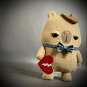 Flat plush sculpture of a capybara with an apathetic expression. It stands on 2 legs like a person and wears a brown beret, a blue bowtie with white polka dots and holds a torn heart.