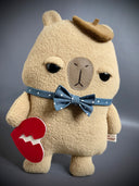 Flat plush sculpture of a capybara with an apathetic expression. It stands on 2 legs like a person and wears a brown beret, a blue bowtie with white polka dots and holds a torn heart.