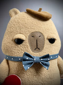Flat plush sculpture of a capybara with an apathetic expression. It stands on 2 legs like a person and wears a brown beret, a blue bowtie with white polka dots and holds a torn heart.