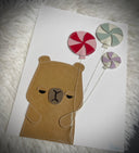 Vinyl fabric art on paper of an unimpressed capybara holding a bunch of pinwheel design round candy balloons.