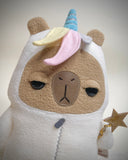 Stuffed animal of a capybara with an unimpressed facial expression and a costume for a white unicorn, with a blue horn and pastel rainbow hair. It holds a star shaped wand.