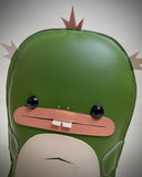 Vinyl fabric plush of a flat dinosaur/kaiju monster with a wide mouth and 2 buckteeth.