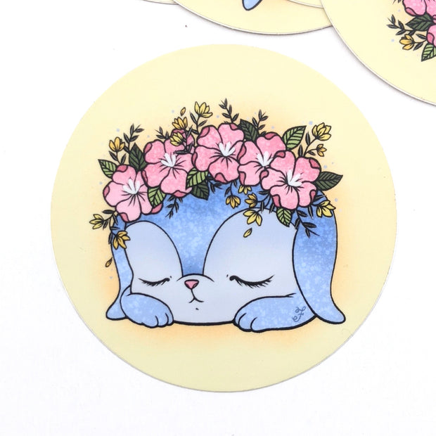Small yellow circular sticker with an illustration of a sleeping blue cartoon bunny with a flower crown atop its head, mostly pink with yellow accent colors.