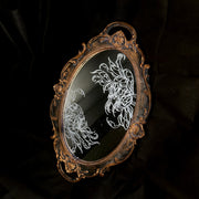 White engraved illustration on a mirror with gold ornate detailing on the exterior. Image is of two flowers, facing one another. The center of one flower is a woman's head and the other is a skeleton's head.