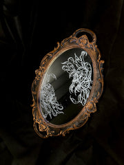 White engraved illustration on a mirror with gold ornate detailing on the exterior. Image is of two flowers, facing one another. The center of one flower is a woman's head and the other is a skeleton's head.