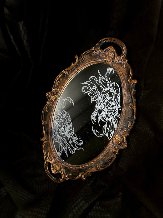 White engraved illustration on a mirror with gold ornate detailing on the exterior. Image is of two flowers, facing one another. The center of one flower is a woman's head and the other is a skeleton's head.