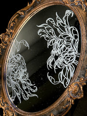 White engraved illustration on a mirror with gold ornate detailing on the exterior. Image is of two flowers, facing one another. The center of one flower is a woman's head and the other is a skeleton's head.