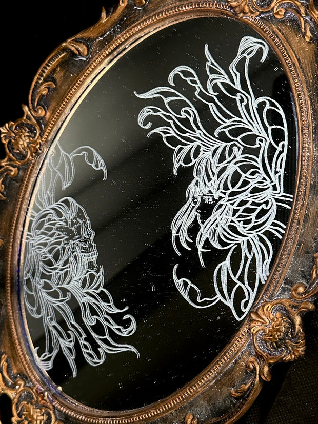 White engraved illustration on a mirror with gold ornate detailing on the exterior. Image is of two flowers, facing one another. The center of one flower is a woman's head and the other is a skeleton's head.