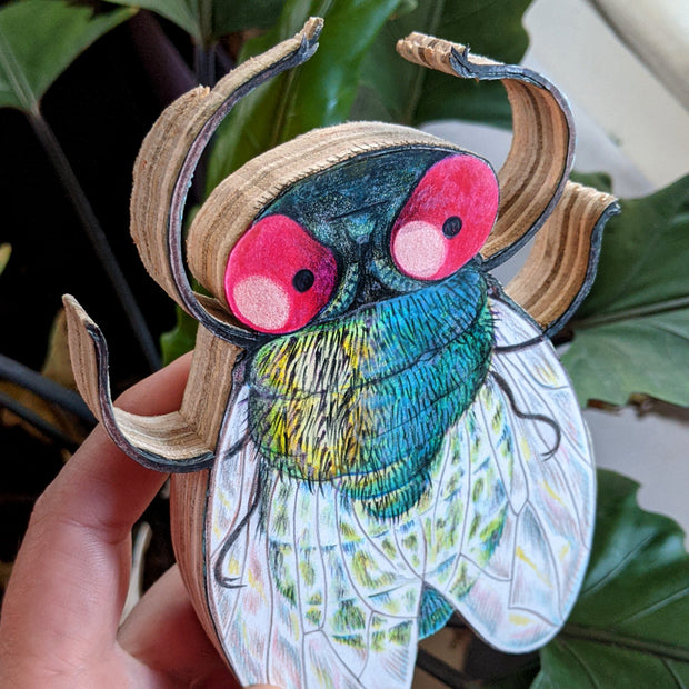 A hand holds a 6.5" fly illustration which is mounted on plywood. The fly has red eyes and a cute, pensive expression. The wings are rainbowy. At this tilted angle, it shows the layers of the plywood.