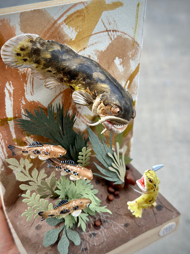 Mixed media diorama style sculpture of a very large, scary fish facing off with a small green frog holding a knife. Green cut paper leaves frame the scene, with small fish swimming nearby.
