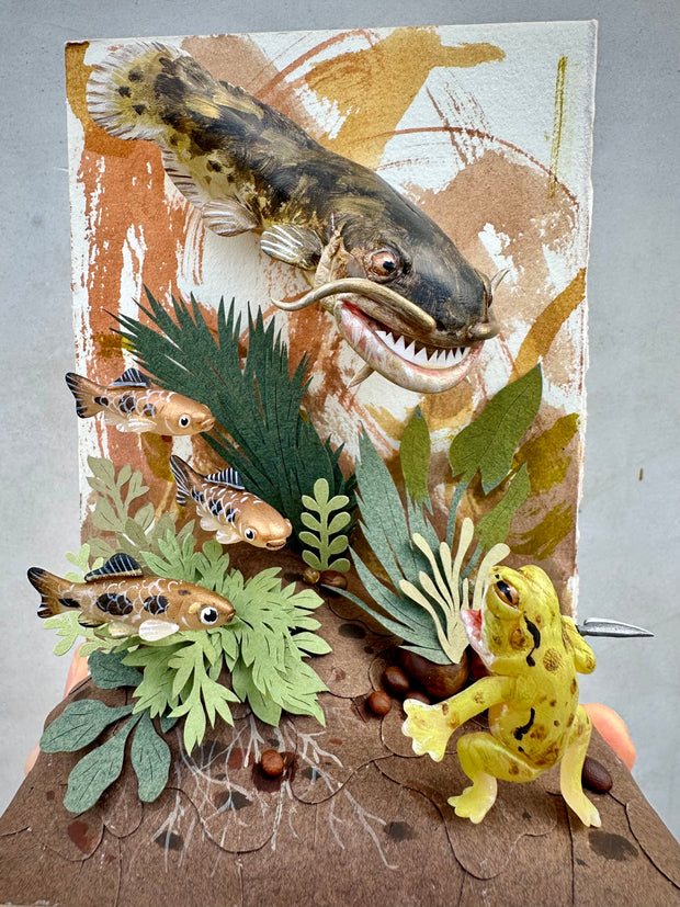 Mixed media diorama style sculpture of a very large, scary fish facing off with a small green frog holding a knife. Green cut paper leaves frame the scene, with small fish swimming nearby.